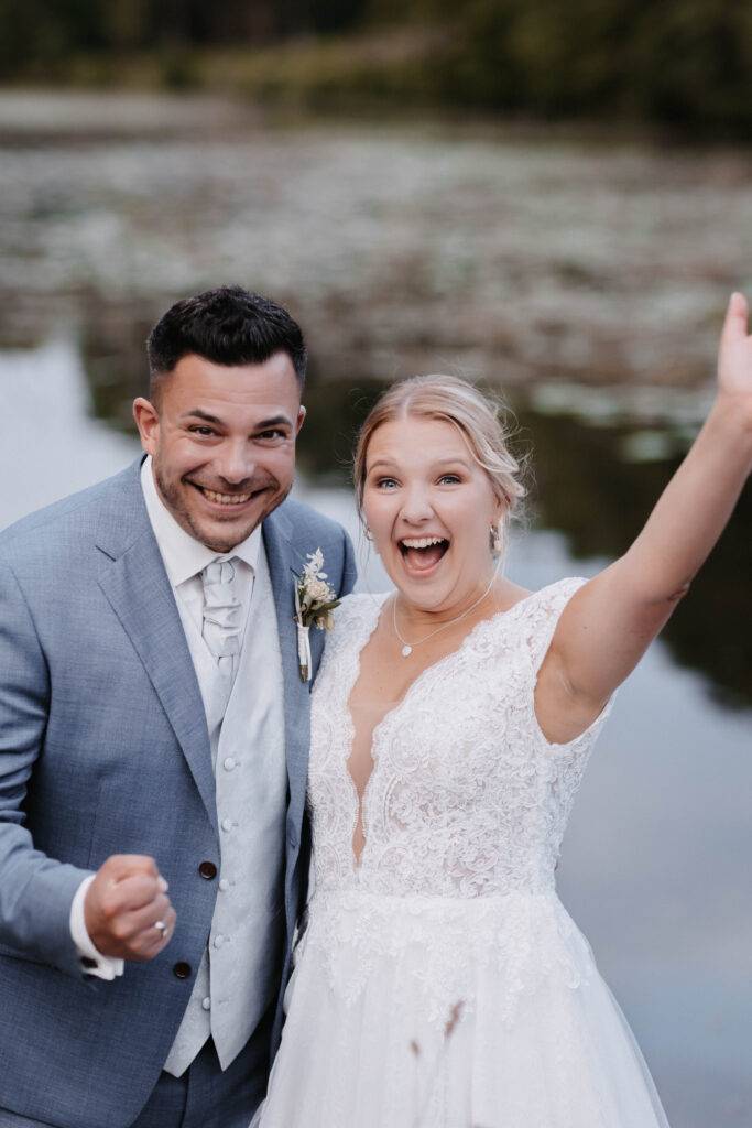 Das Brautpaar schaut mit einem fröhlichen Gesicht in die Kamera und steht bei einem See.Hochzeitsfotograf-Hamburg