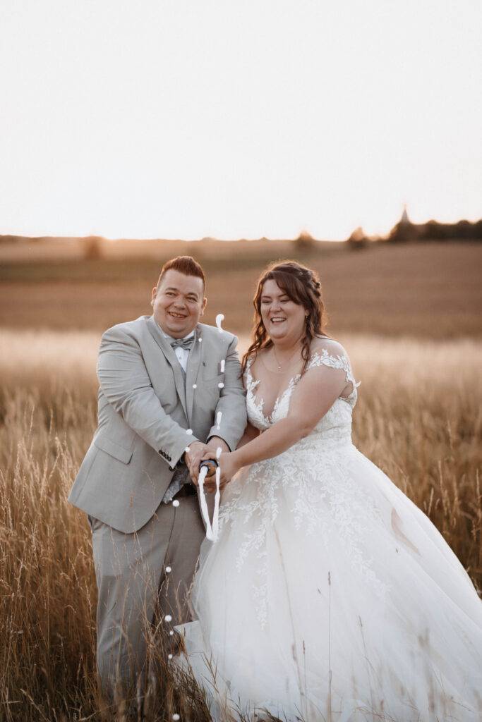 Das Brautpaar schüttelt eine Sektflasche bei Sonnenuntergang in einem Weizenfeld. Hochzeitsfotograf-Hamburg