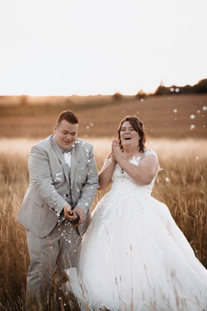 Das Brautpaar schüttelt eine Sektflasche bei Sonnenuntergang in einem Weizenfeld. Hochzeitsfotograf-Hamburg