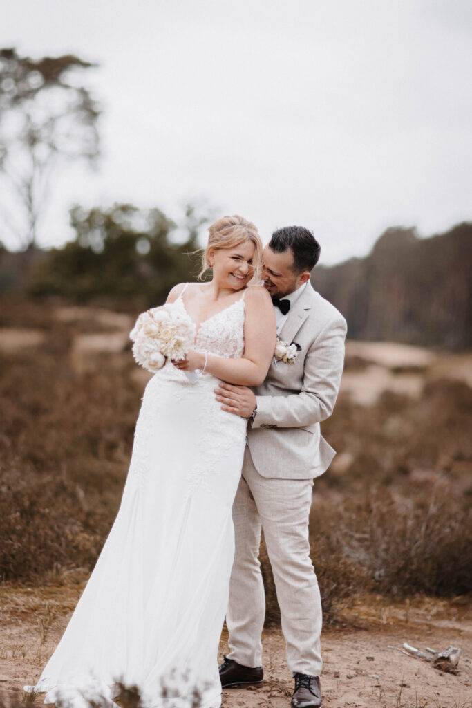 Das Brautpaar beim Brautpaarshooting. Sie steht vor ihm und er umarmt sie von hinten. Sie hält ihren Brautstrauß in beiden Händen. Hochzeitsfotograf-hamburg