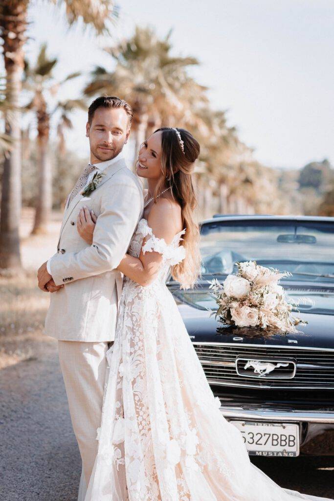 Das Brautpaar steht in einer Allee von Palmen hintereinander und schauen sich dabei an. Hinter ihnen steht ein Mustang mit Blumen beschmückt. Hochzeitsfotograf-Hamburg