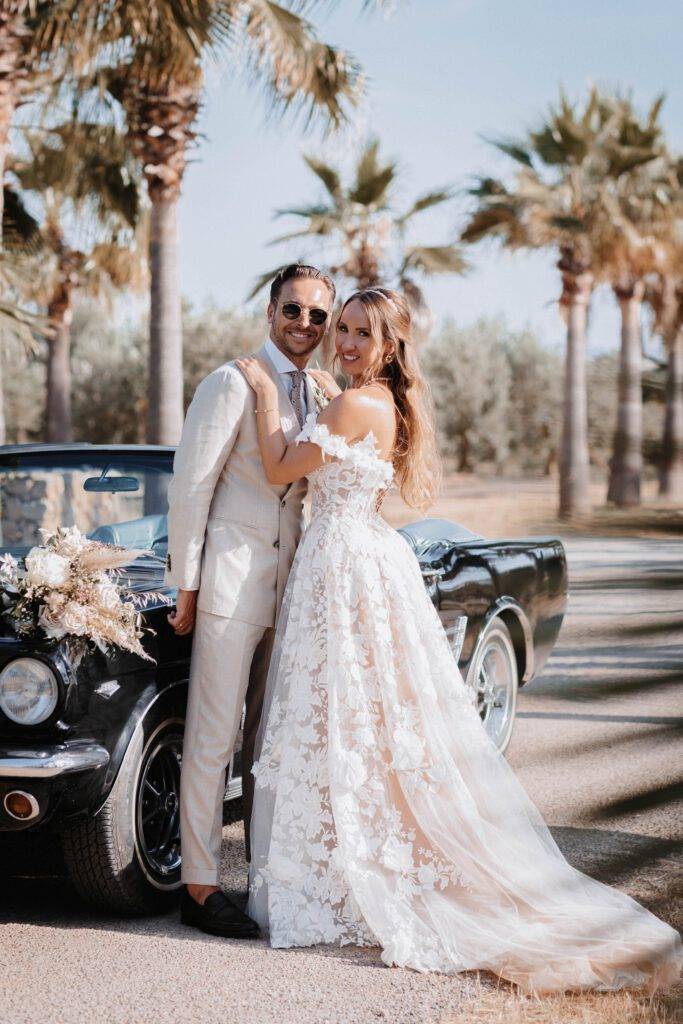 Das Brautpaar steht in einer Allee von Palmen hintereinander und schauen sich dabei an. Hinter ihnen steht ein Mustang mit Blumen beschmückt. Hochzeitsfotograf-Hamburg