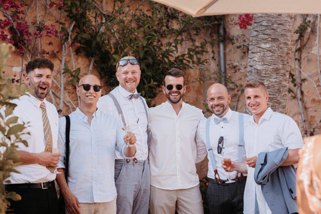 Gruppenaufnahme von Gästen auf einer Hochzeit auf Mallorca. Alle schauen in die Kamera und halten dabei jeweils ein Getränk in der Hand
