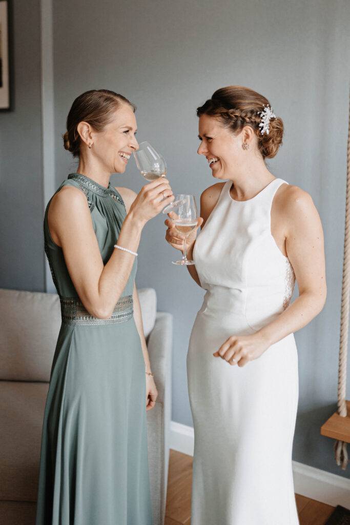 Die Braut steht steht mir ihrer Trauzeugin voreinander und beide halten ein Glas Sekt in der Hand. Beide lächeln. Die Braut hat ein weißes enganliegendes Kleid an und die Trauzeugin ein grünes. Hochzeitsfotograf-hamburg