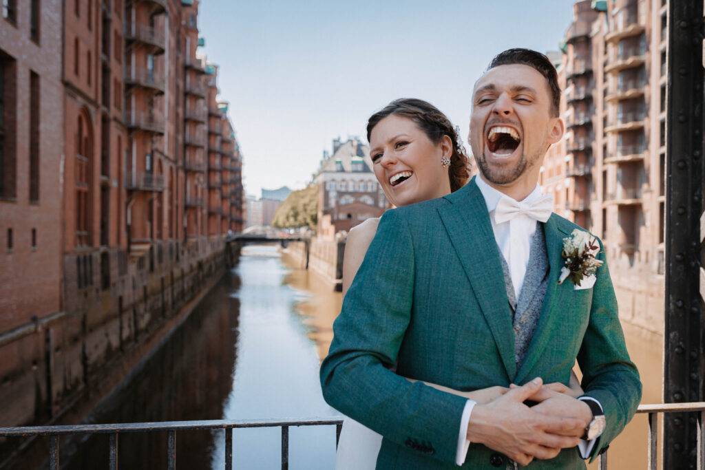 Das Brautpaar paar steht voreinander und lacht. Sie hat ihren Mann von hinten Umarmt. Im Hintergrund ist wasser zu sehen. Hochzeitsfotograf-Hamburg