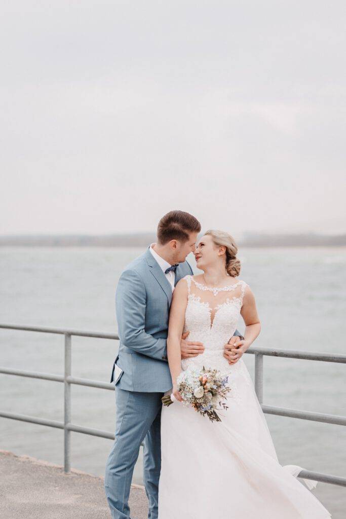 Das Brautpaar steht am Meer voreinander und Küssen sich dabei. Der Bräutigam hat seine Braut von hinten im Arm.