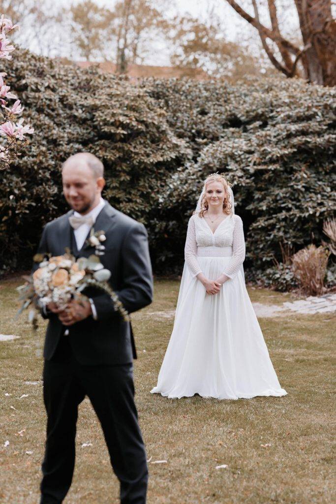 First Look.´Der Bräutigam steht mit dem Rücken zur Braut im Vordergrund und die Braut steht hinter ihn und wartet darauf, dass er sie das erstemal im Brautkleid sieht