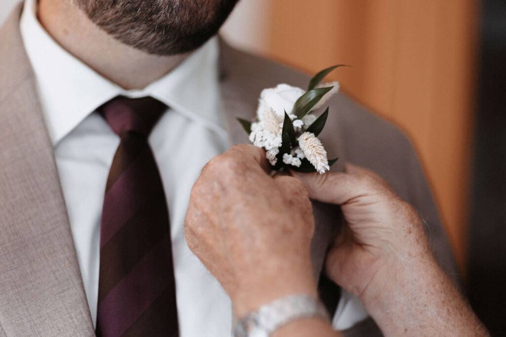 Zu sehen ist eine Detailaufnahme, wie eine weiße Blume an den Anzug eines Mannes gesteckt wird.