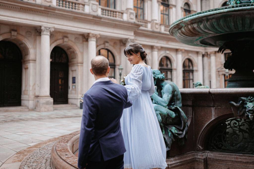 Ein junges, festlich gekleidetes Pärchen hält sich an der Hand und spaziert um den Brunnen des Hamburger Rathauses.