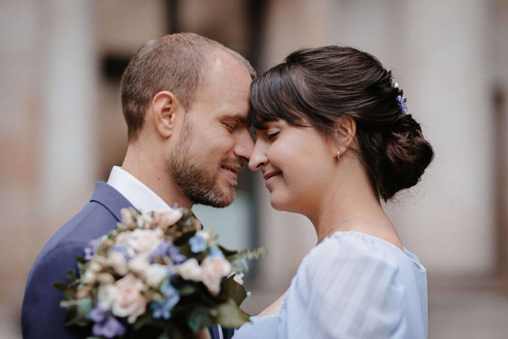 Eine Nahaufnahme von einem festlich gekleideten Pärchen, beide schmiegen die Stirn aneinander. Die Frau hält einen Strauß in der Hand.