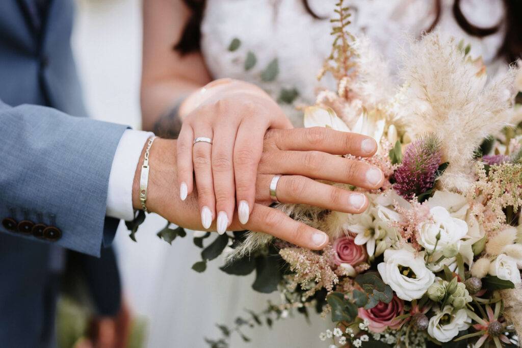Eine Deatilaufnahme von zwei Händen, an denen Eheringe stecken. Im Hintergrund ist ein Bkumenstrauß zu sehen.