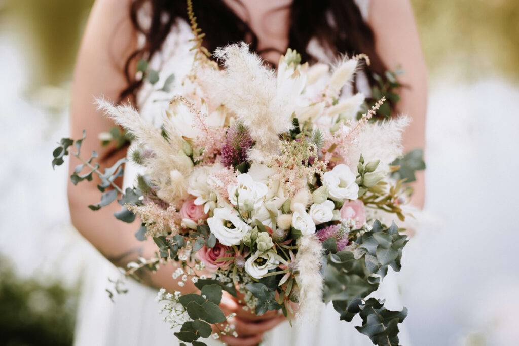 Zu sehen ist ein bunter Blumenstrauß. Im Hintergrund ist unscharf eine Braut zu erkennen.