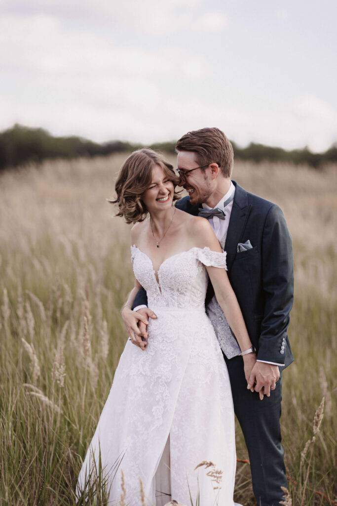 Ein junges Ehepaar steht in mitten von hohen Gräsern, hält sich an den Händen und beide lachen.