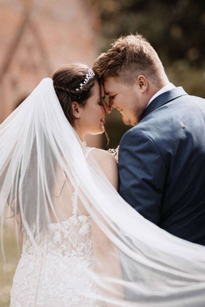 Stirn an Stirn steht das Brautpaar voreinander unter dem Schleier der Braut