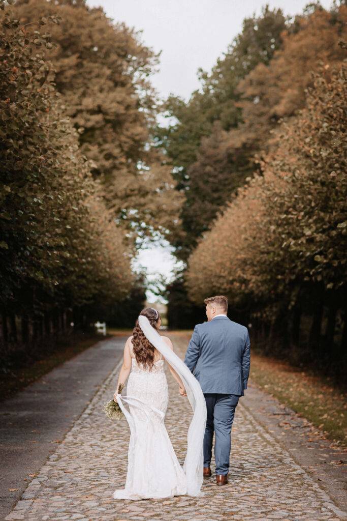 Das Brautpaar geht mittig in einer Allee und schaut sich dabei an