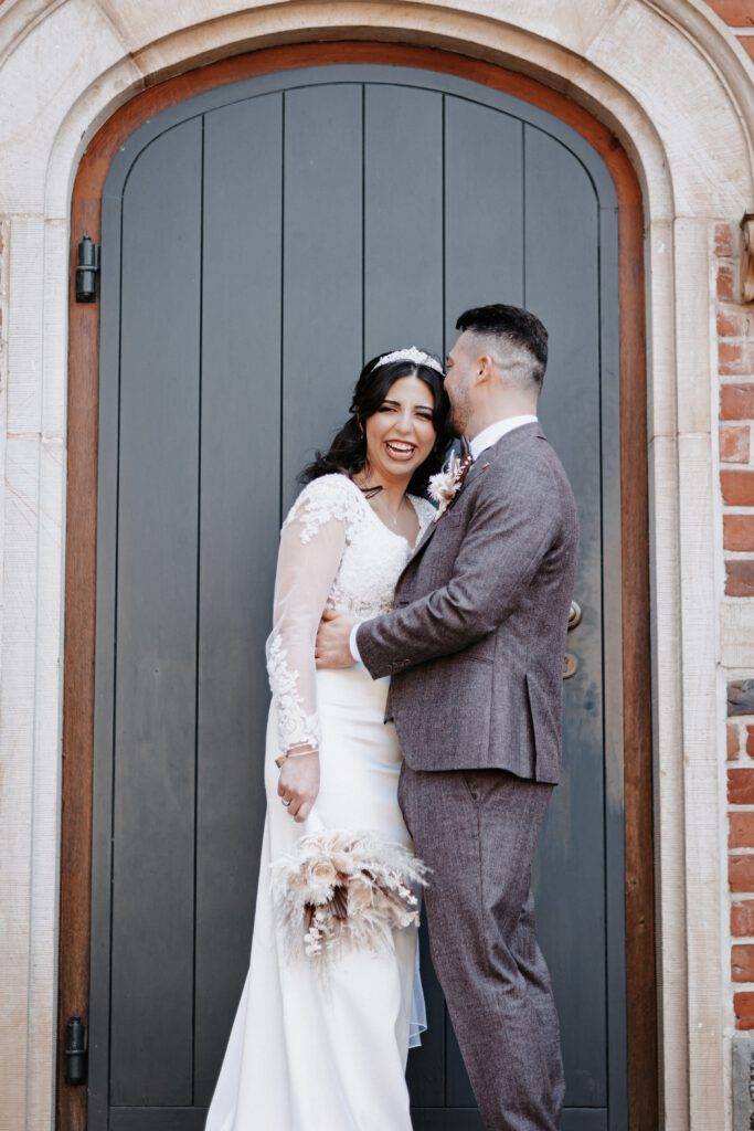 Vor einer Tür steht das Brautpaar. Die Braut schaut lachend n die Kamera während der Bräutigam Sie seitlich am Kopf küsst. Hochzeitsfotograf Hamburg. Hochzeitsfilm Hamburg