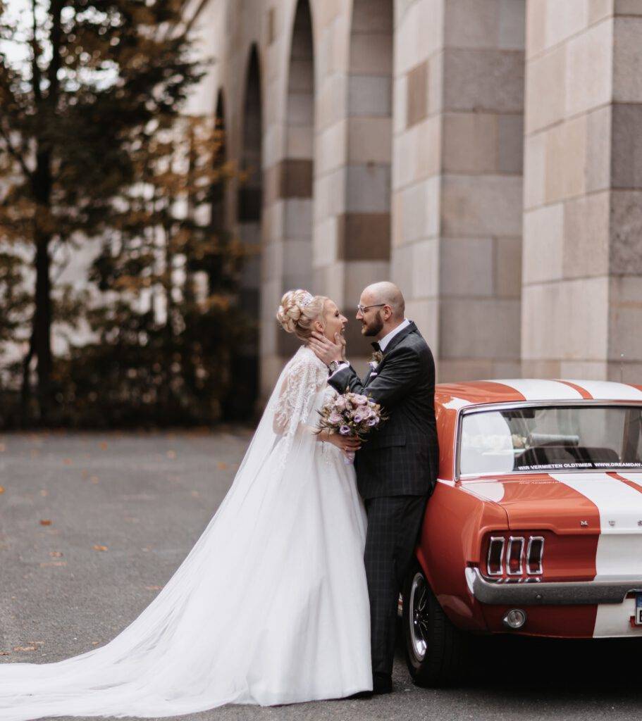 Das Bruatpaar steht vor einander. Der Bräutigam ist dabei vor einem roten alten Mustang angelehnt. Hochzeitsfotograf Hamburg. Hochzeitsfilm Hamburg