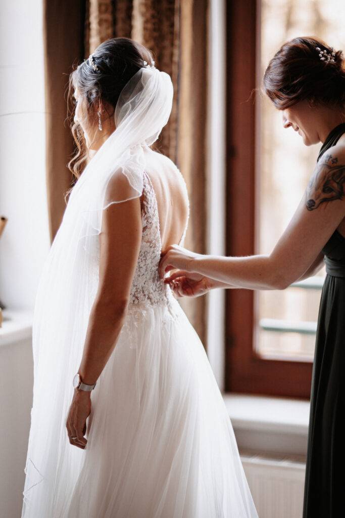 Die Trauzeugin schließt das Kleid mit dem Reißverschluss vom Rücken des Hochzeitskleides der Braut.Hochzeitsfotograf Hamburg. Hochzeitsfilm Hamburg