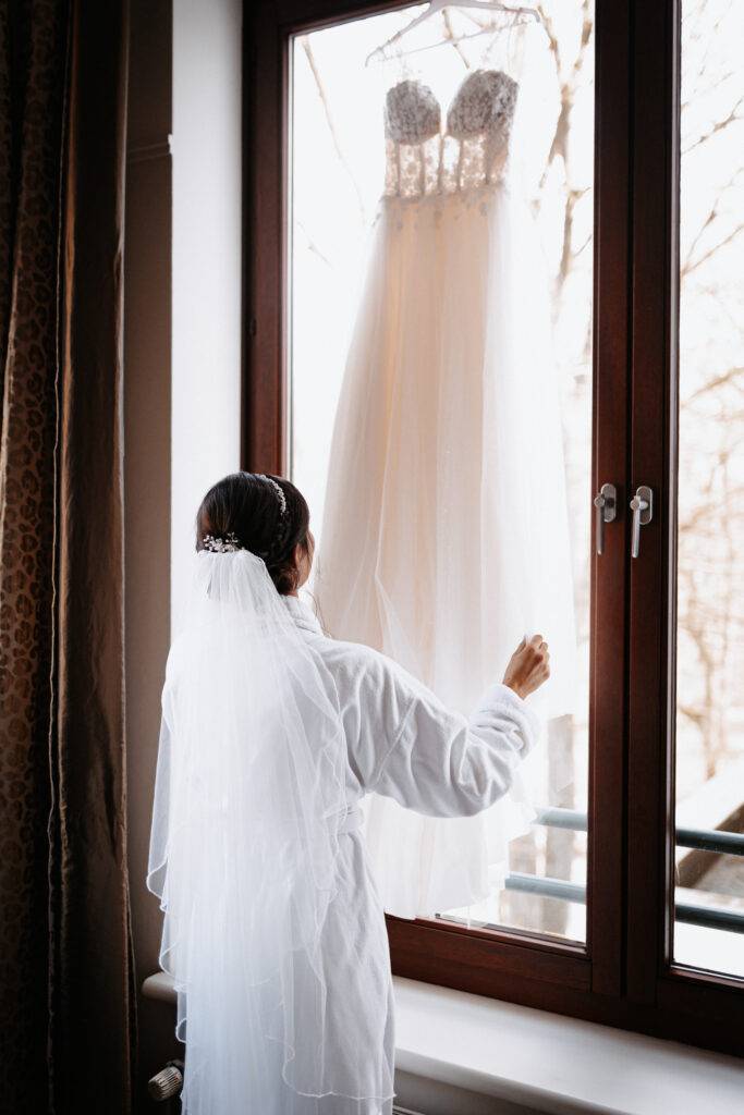 Die Braut steht vor ihrem Hochzeitskleid, welches vor dem Fenster aufgehängt ist und schaut es an. Hochzeitsfotograf Hamburg. Hochzeitsfilm Hamburg