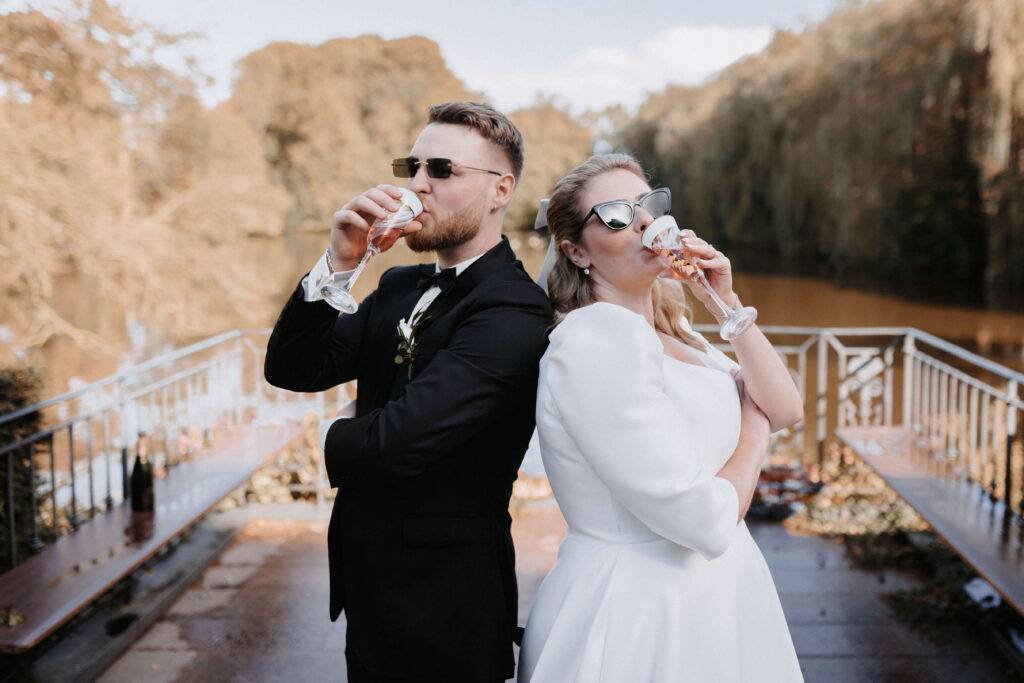 Das Hochzeitspaar trinkt aus einem Sektglas und haben Sonnenbrillen auf